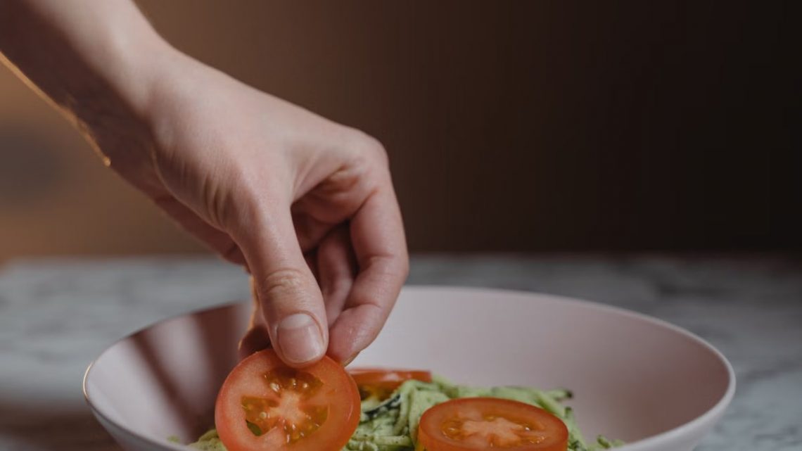 A close-up photo of a hand placing fresh tomato slices on a vegetable salad in a bowl.