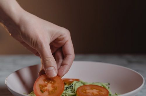 A close-up photo of a hand placing fresh tomato slices on a vegetable salad in a bowl.