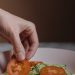 A close-up photo of a hand placing fresh tomato slices on a vegetable salad in a bowl.